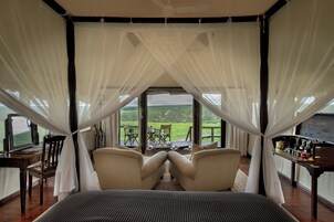 A bedroom with a large bed, two chairs, a small table, and a view of a green landscape through large windows.