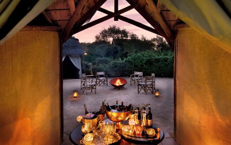 A cozy tented area with a table set for dining, surrounded by chairs and a fire pit, under a thatched roof.