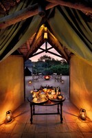 A cozy tented area with a table set for dining, surrounded by chairs and a fire pit, under a thatched roof.