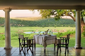 A covered outdoor dining area with a table set for two, overlooking a grassy landscape with zebras. - Gorah Elephant Camp (Addo)