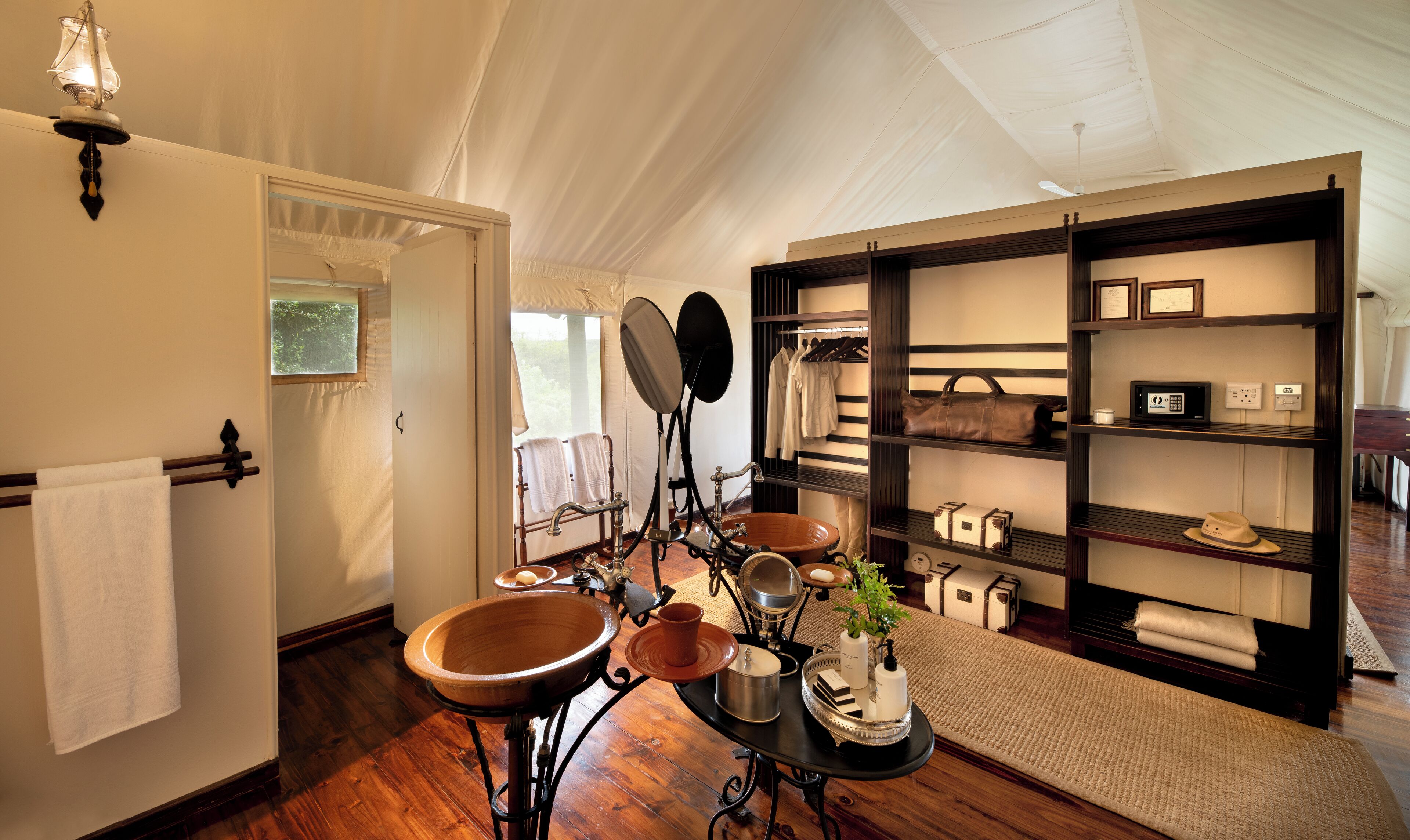 A tented room with a wooden floor, a large wooden basin, a black metal table with toiletries, and a shelf with bags and a hat.
