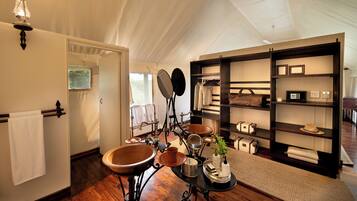 A tented room with a wooden floor, a large wooden basin, a black metal table with toiletries, and a shelf with bags and a hat.