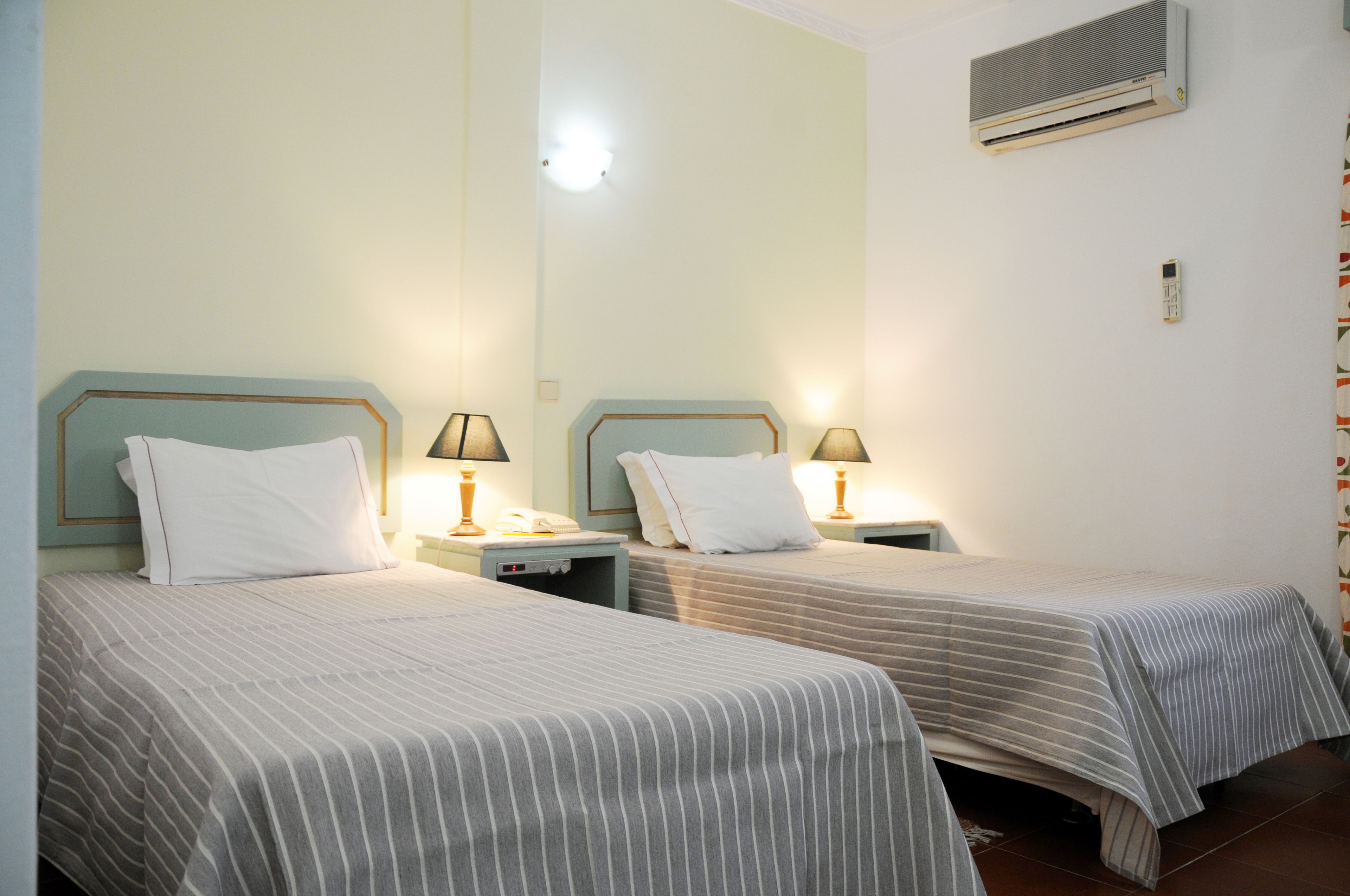 Two single beds with striped bedding, bedside lamps, and a wall-mounted air conditioner.