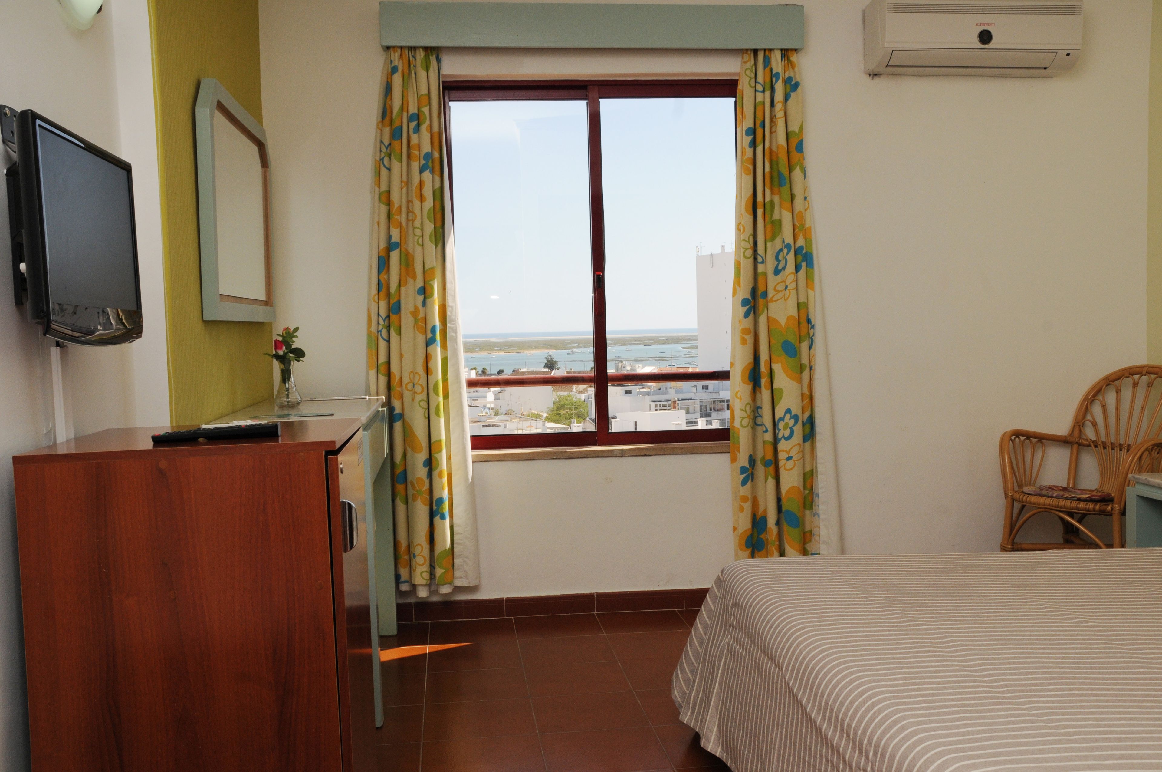 A bedroom with a bed, a desk, a chair, a TV, and a window with curtains.