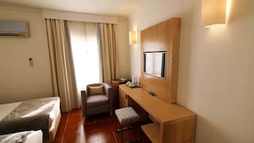 A hotel room with two beds, a desk, a chair, a small stool, a TV, and a window with curtains.