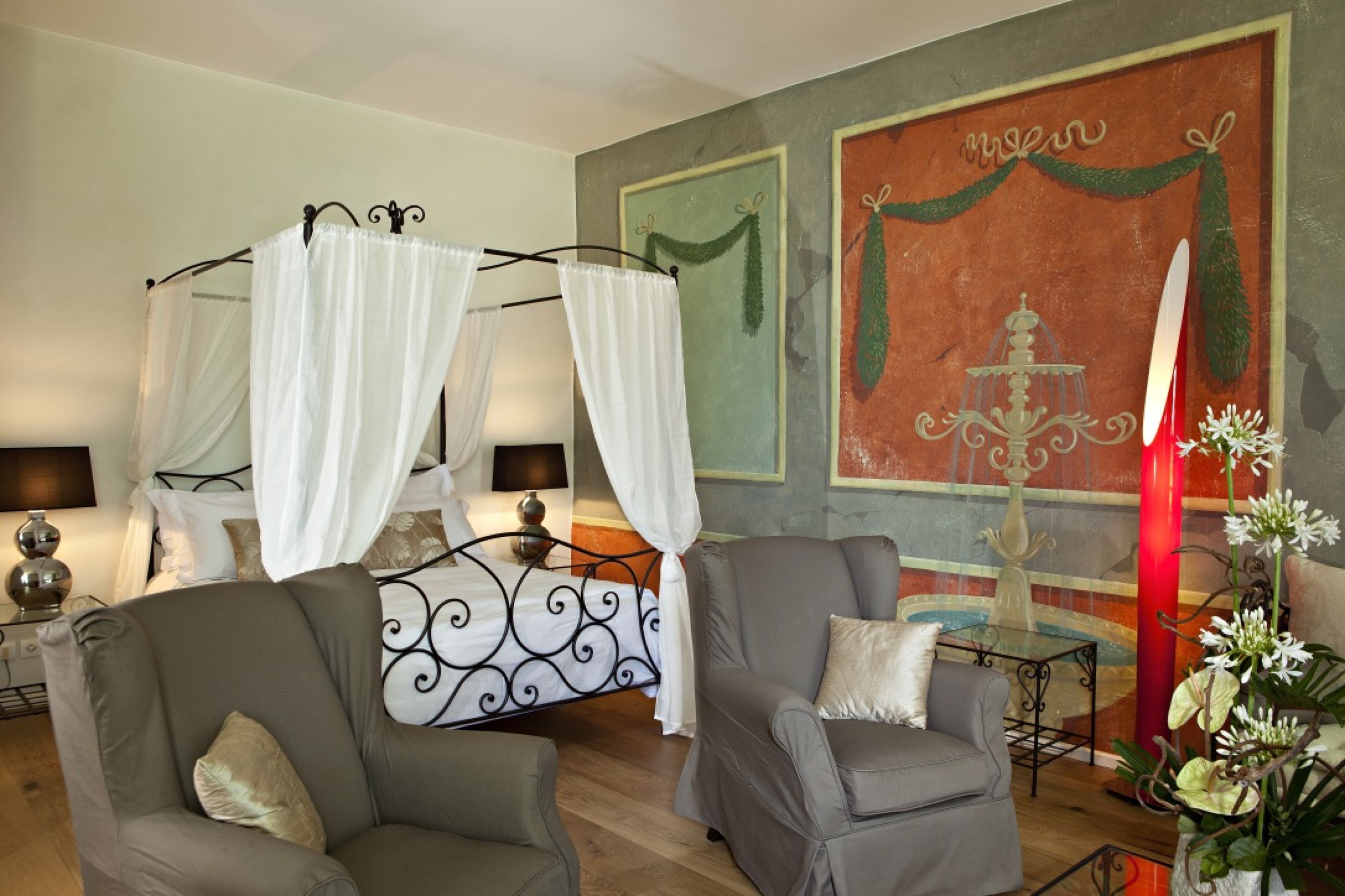 Une chambre à coucher avec un lit à baldaquin, deux fauteuils, une table de chevet, une lampe et un mur décoratif agrémenté d’un lustre.
