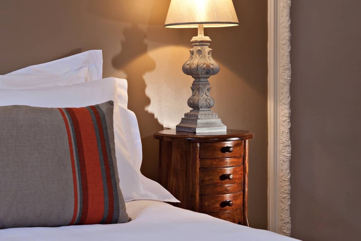 A neatly made bed with white linens and a striped pillow, a wooden nightstand with a lamp, and a wall with a decorative molding.