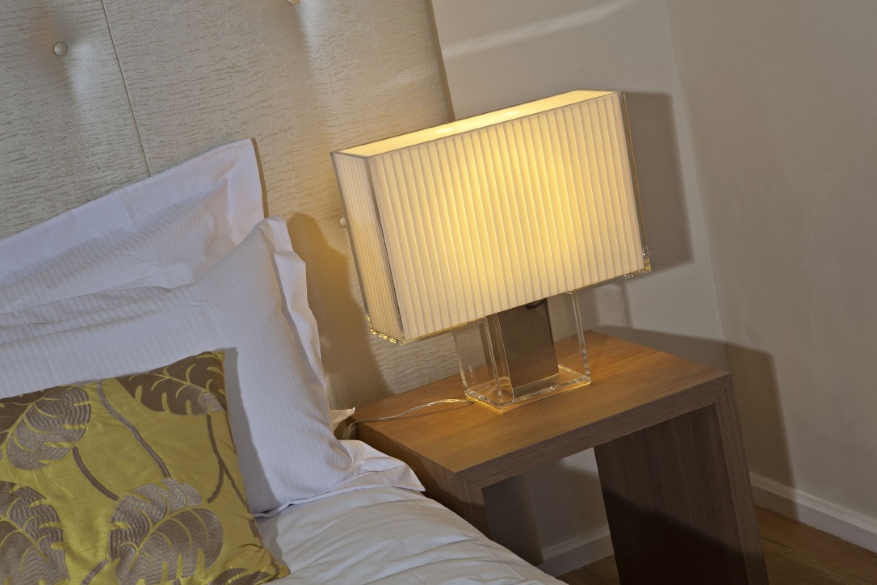 Una cama bien hecha con sábanas blancas y un cojín amarillo con estampado de hojas. Se ve una mesita de noche con una lámpara de pie y un mueble de madera.