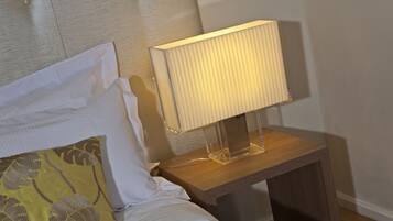 Una cama bien hecha con sábanas blancas y un cojín amarillo con estampado de hojas. Se ve una mesita de noche con una lámpara de pie y un mueble de madera.