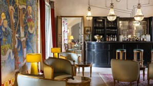 Una zona de bar con techo de madera, candelabros colgantes y una barra repleta de botellas. Se ven varias sillas y una mesa en primer plano.