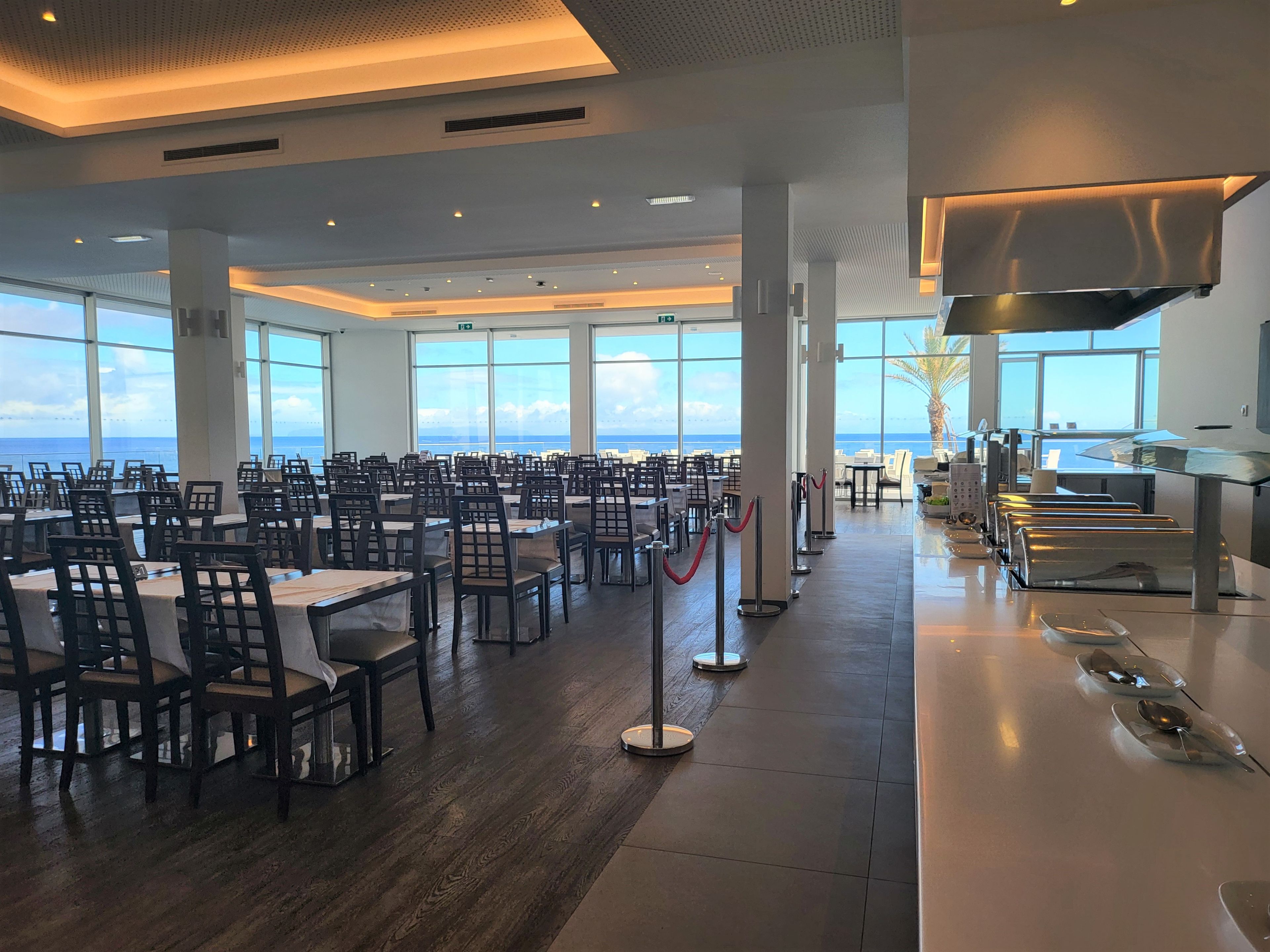 Un vaste espace de salle à manger avec des tables et des chaises, de grandes fenêtres offrant une vue sur l’océan, et une cuisine moderne équipée d’appareils électroménagers en acier inoxydable.