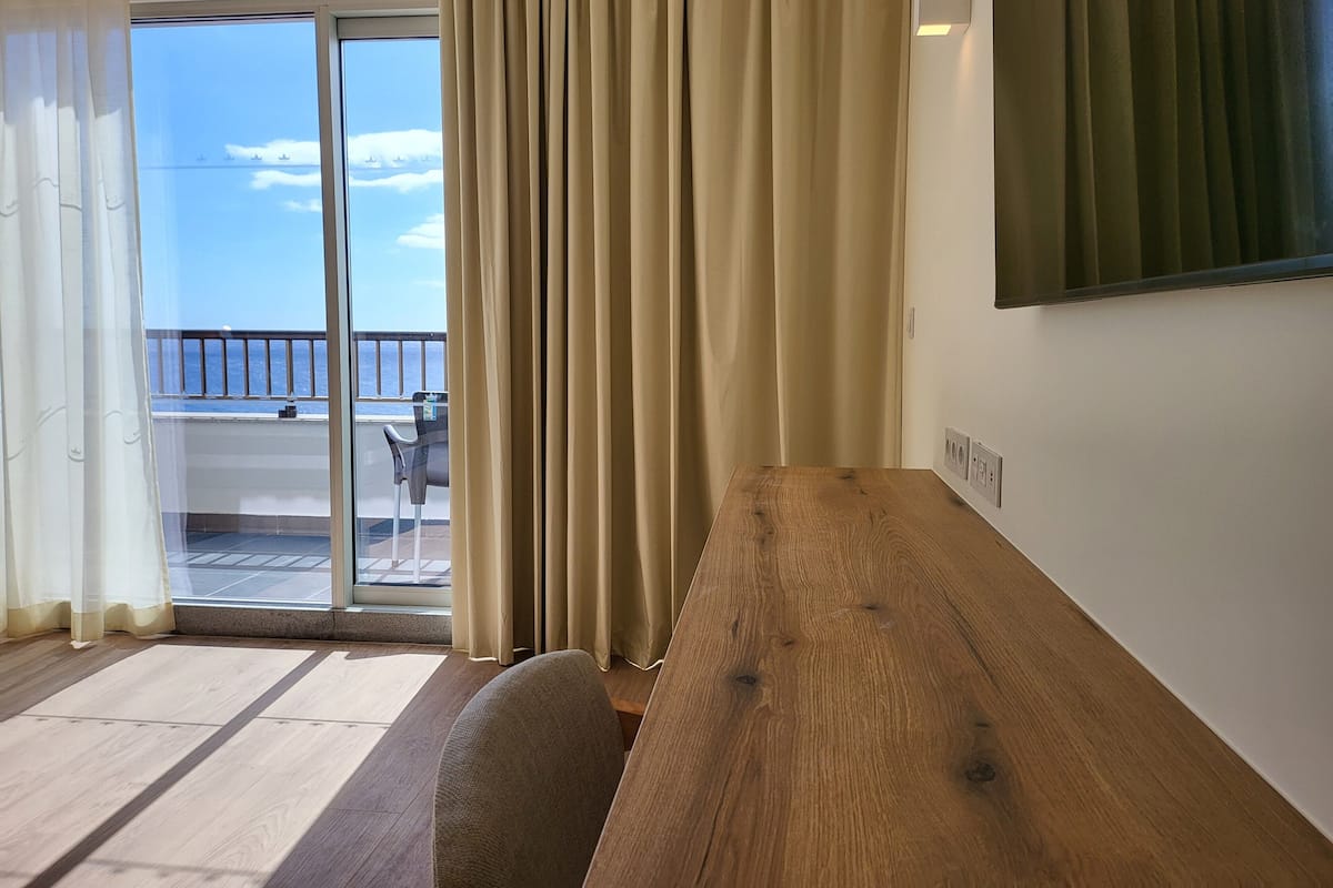 A room with a wooden bench, a sliding glass door leading to a balcony with a view of the sea, and a wall-mounted TV.