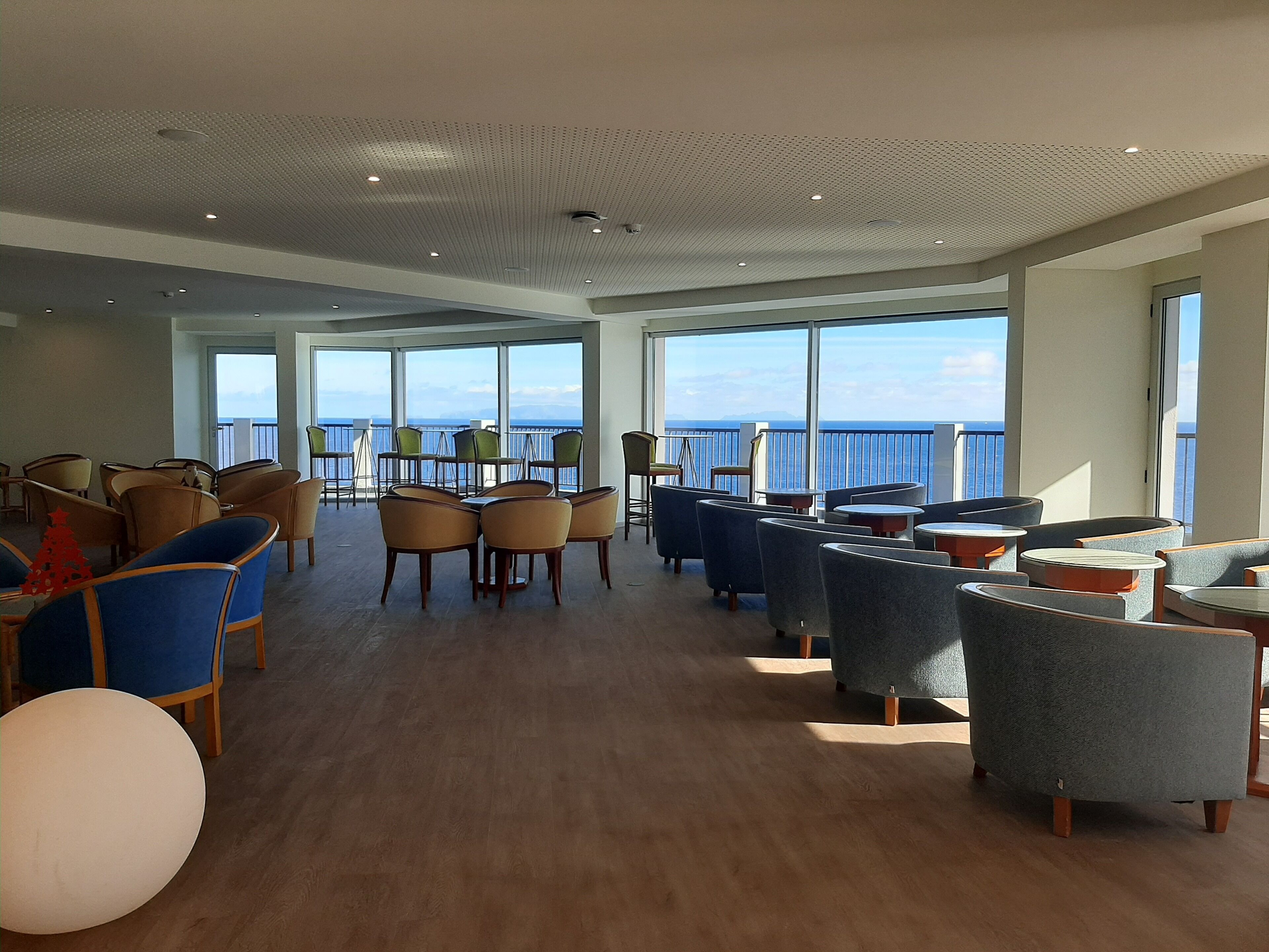 Een ruime loungeruimte met diverse zitplaatsen, grote ramen met uitzicht op zee, en een hoog plafond met inbouwverlichting.