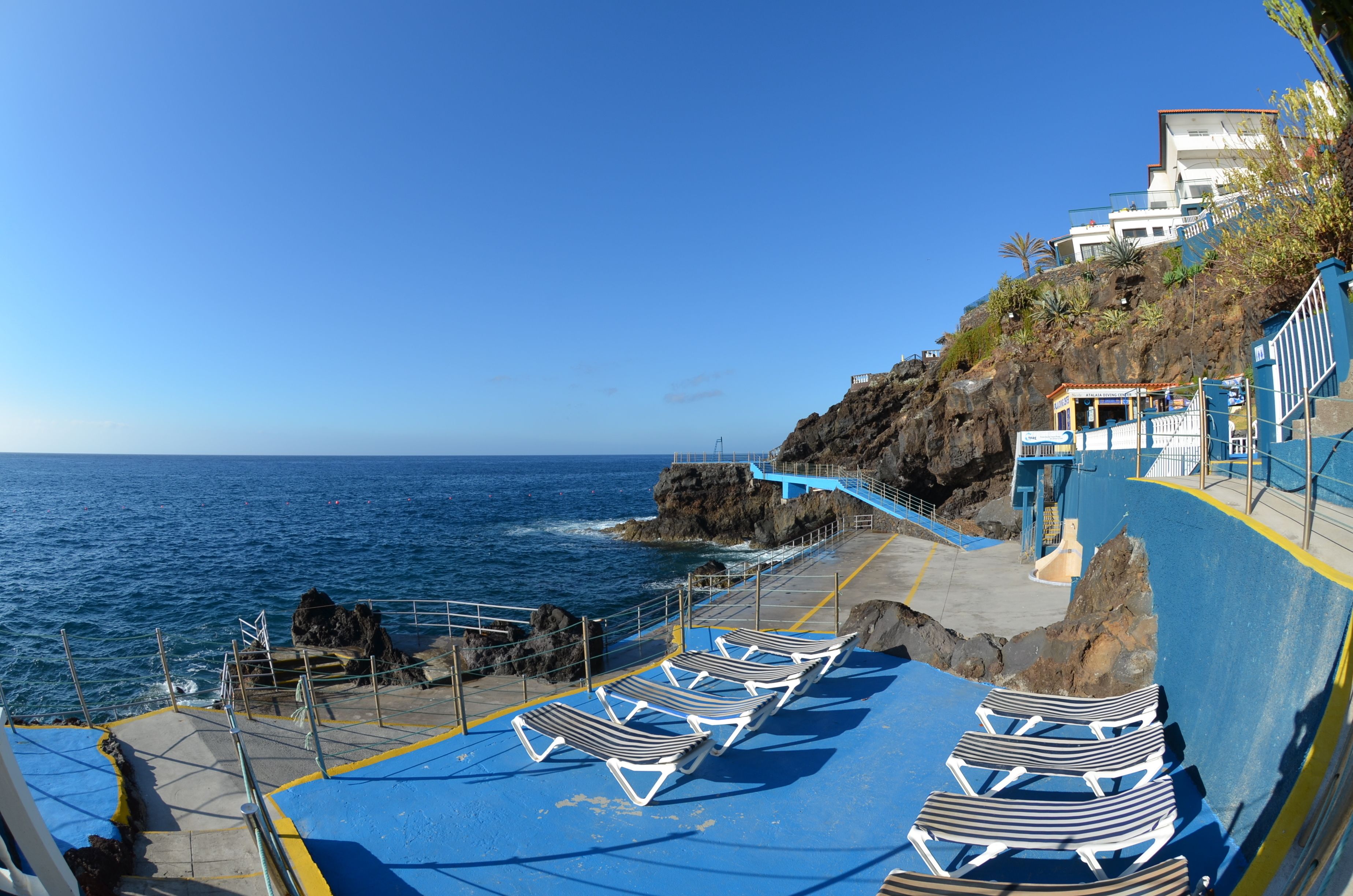 A coastal area with a rocky cliff, a blue walkway, and lounge chairs by the sea.