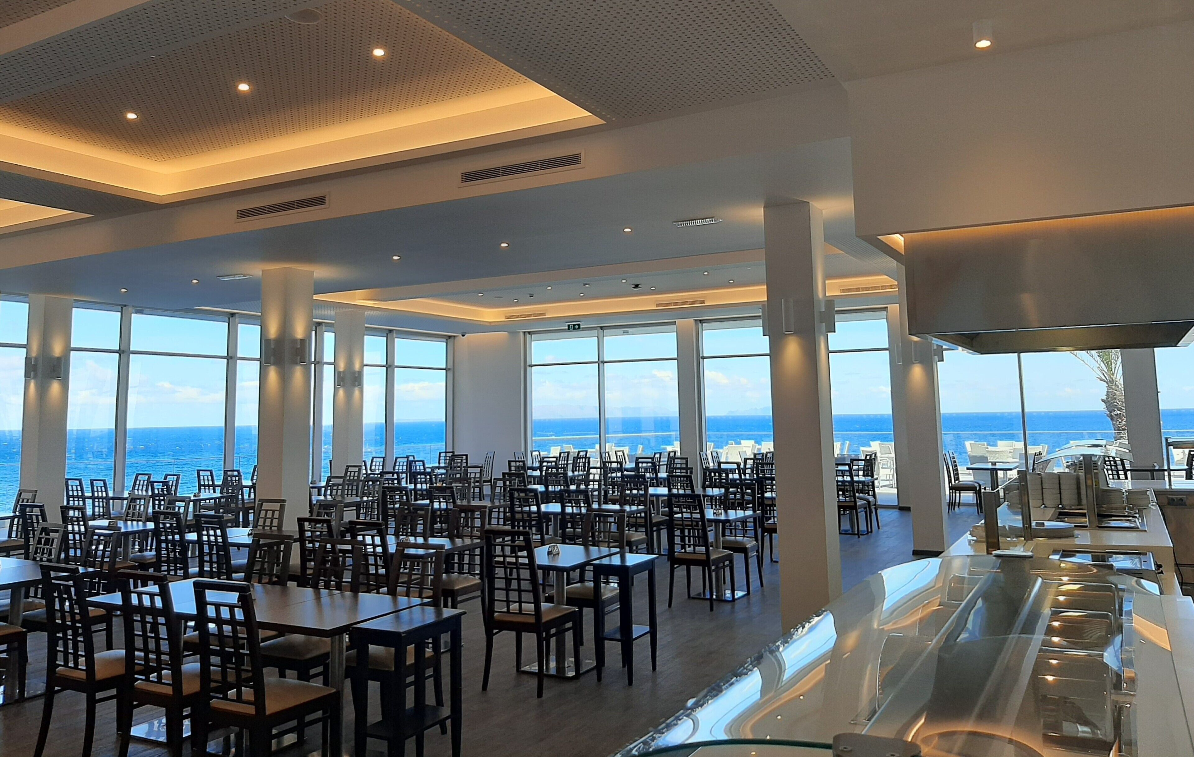 A spacious dining area with tables and chairs, large windows offering a view of the ocean, and a modern ceiling design.
