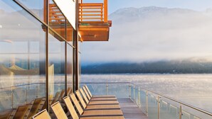Terraza cubierta con paredes de vidrio, tumbonas de madera y barandilla de vidrio, con vista a un lago helado y montañas.