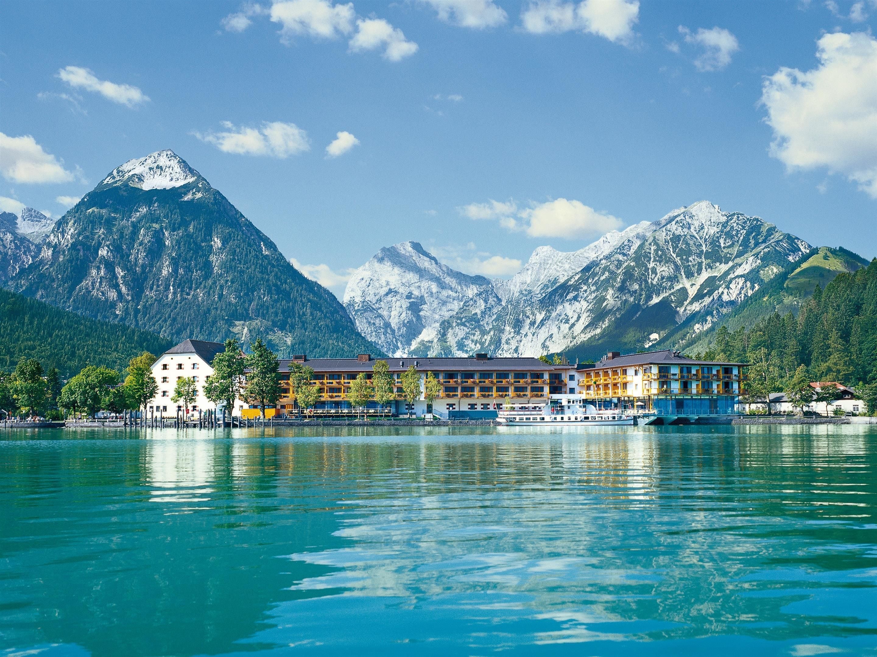 A hotel with multiple floors and balconies, situated by a lake with mountains in the background.