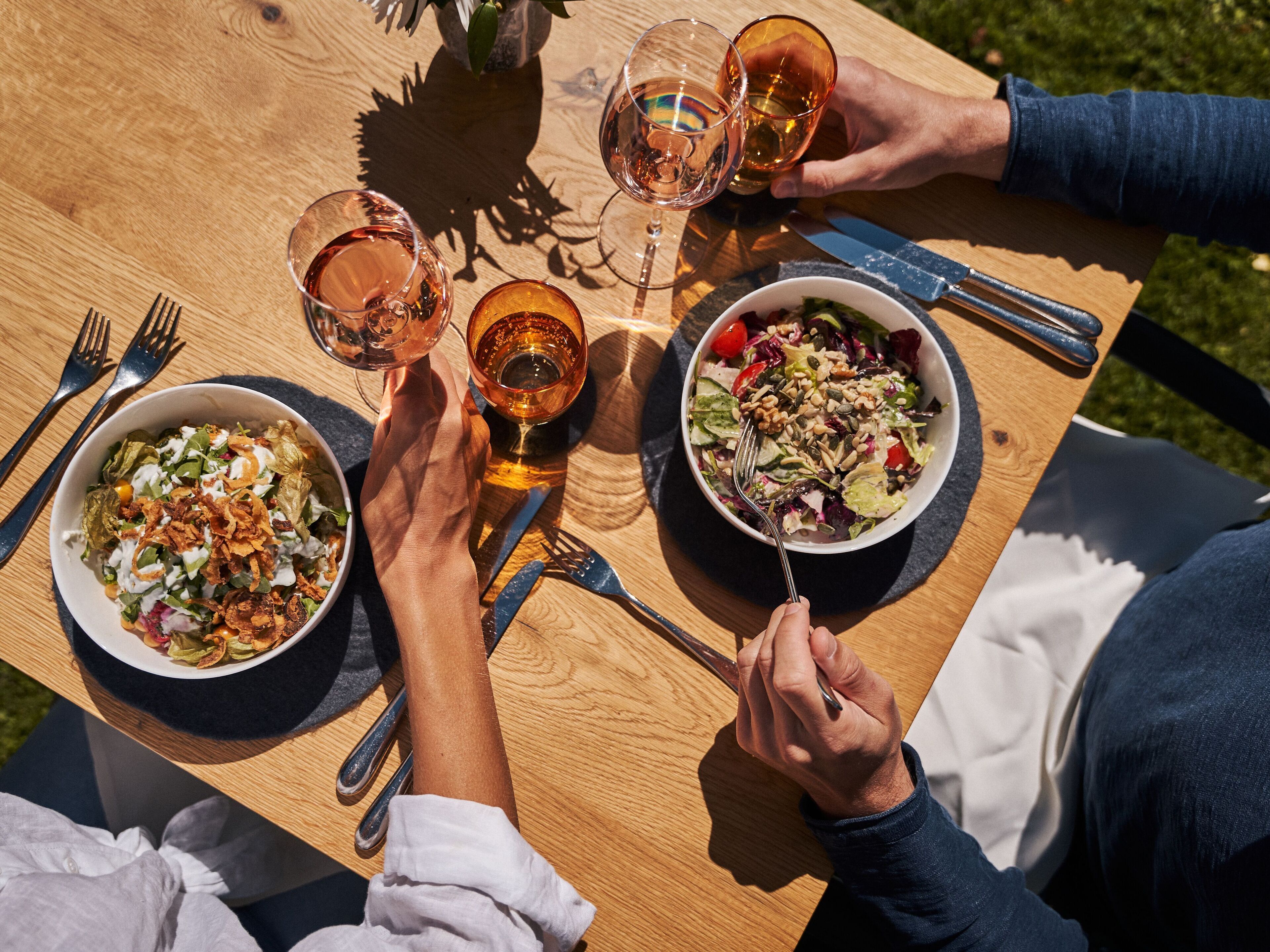 A wooden table with two bowls of food and two glasses of amber liquid, likely wine, with forks and knives on the table.