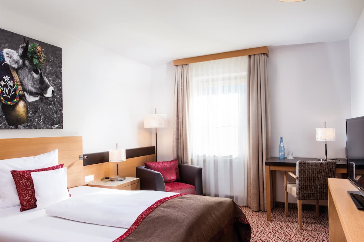 Une chambre d’hôtel avec un lit, deux chaises, un bureau, une télévision et une fenêtre avec des rideaux.