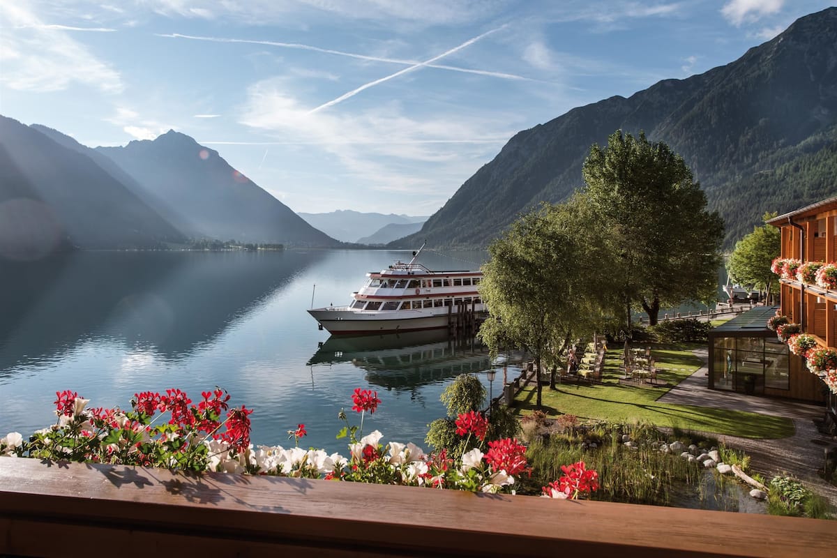 Un lac où est amarré un bateau, entouré de montagnes et bordé d’un jardin bien entretenu, agrémenté de fleurs.