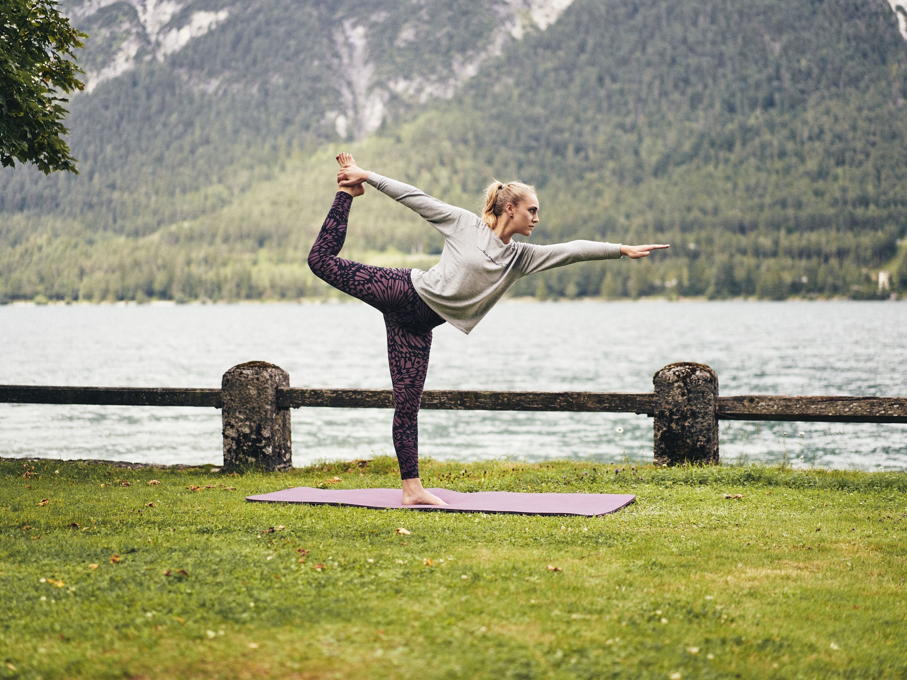 A person performing a yoga pose on a mat in a grassy area with a lake and mountains in the background.
