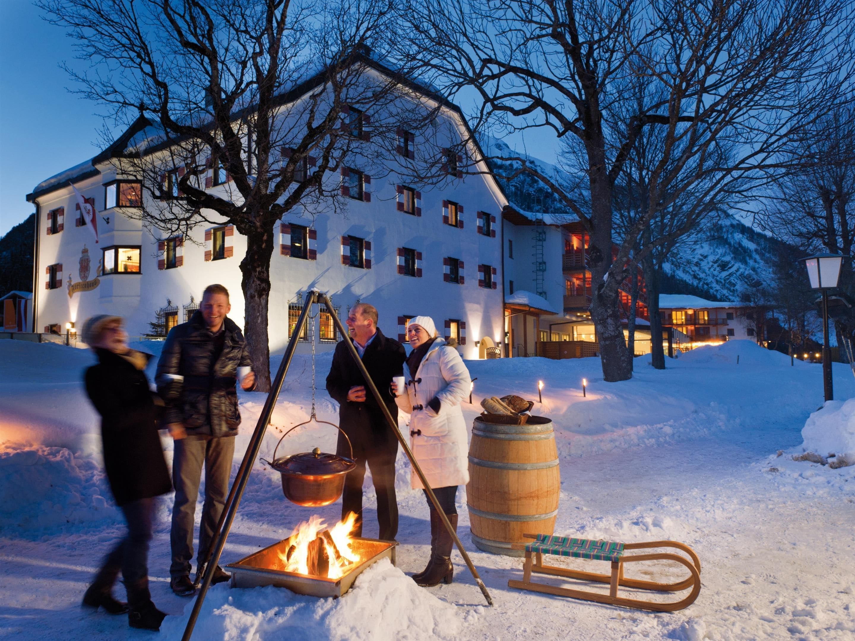 A group of people gathered around a campfire in a snowy outdoor setting with a large building in the background.