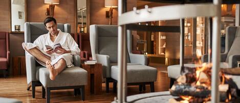 A man reading in a modern lounge with grey armchairs, a fireplace, and wooden flooring.