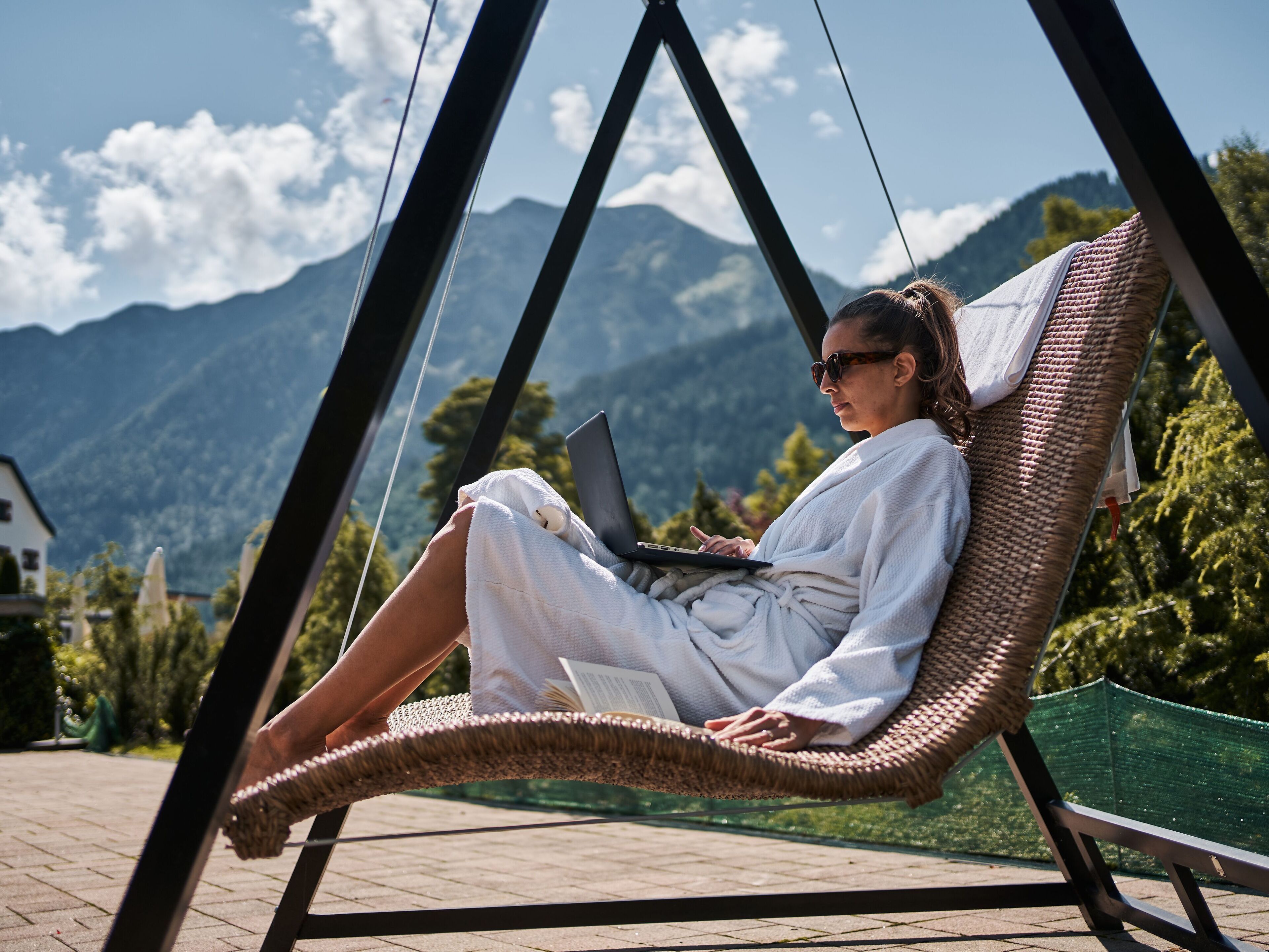 A person relaxing in a wicker swing chair with a laptop, overlooking a mountainous landscape.