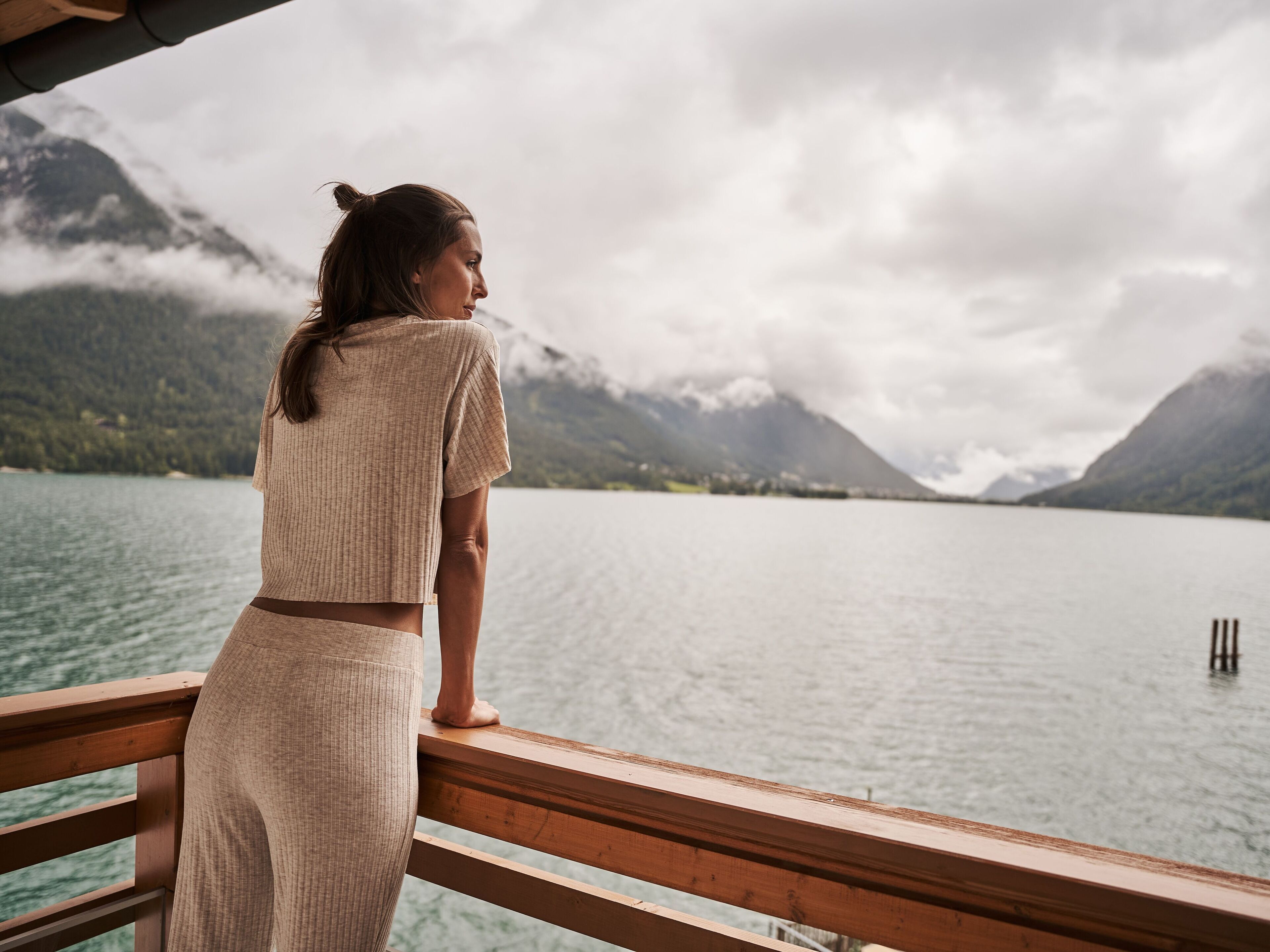 A person standing on a wooden deck overlooking a calm lake with mountains in the background.