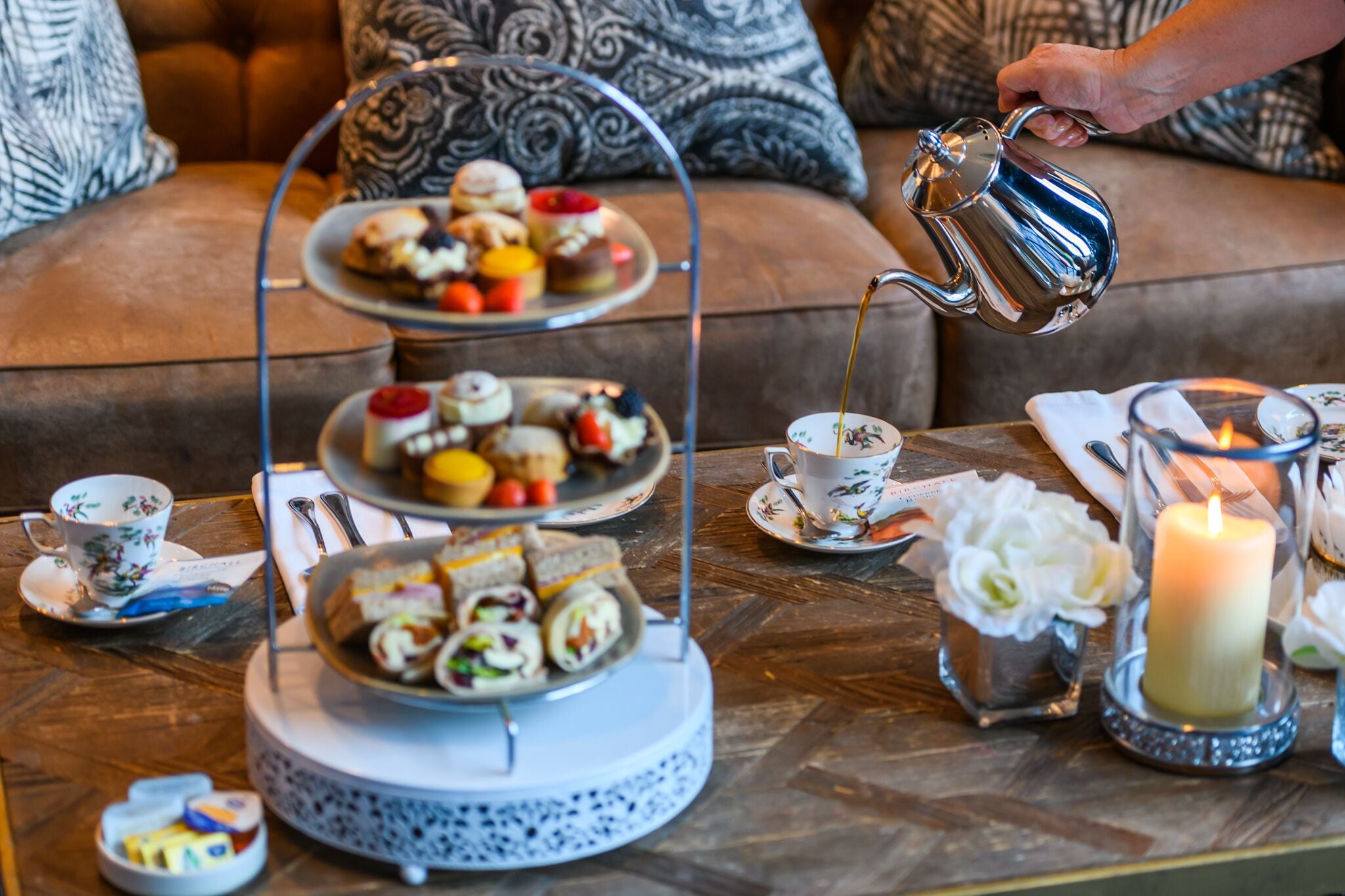 A three-tiered dessert stand with various pastries and sandwiches, a teapot pouring tea, a candle, and a floral arrangement on a wooden table.
