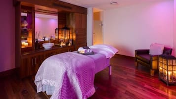 A spa room with a wooden cabinet, a bed with a white and purple blanket, a chair, and a lantern.