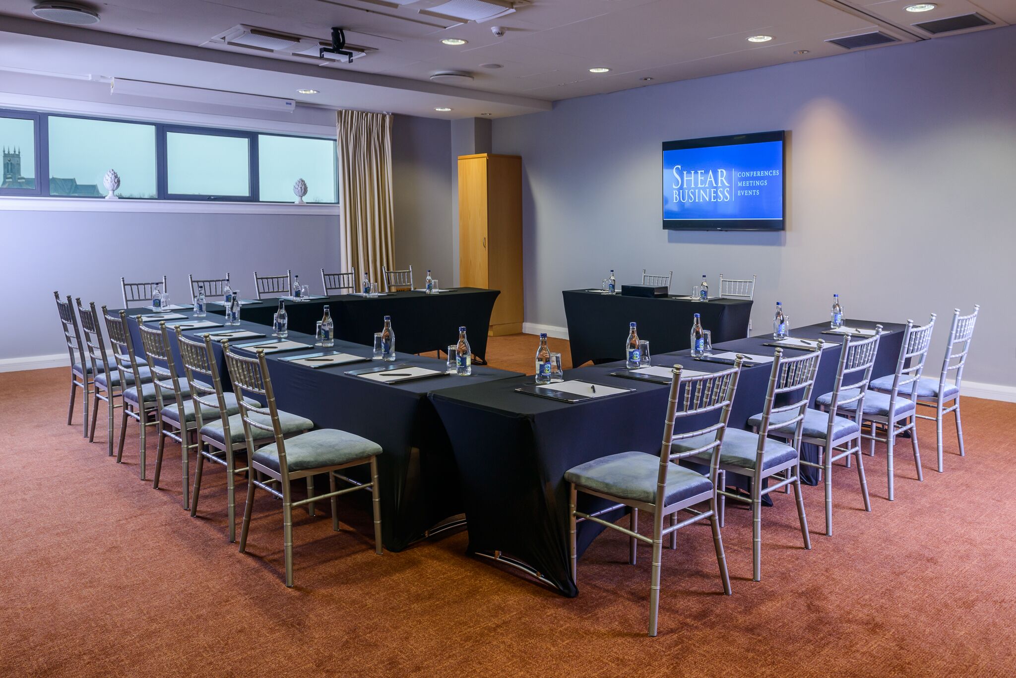 Una sala conferenze con diversi tavoli e sedie, dotata di bottiglie d'acqua e cartellini segnaposto, e uno schermo che mostra "Shear Conferences Meetings Events".