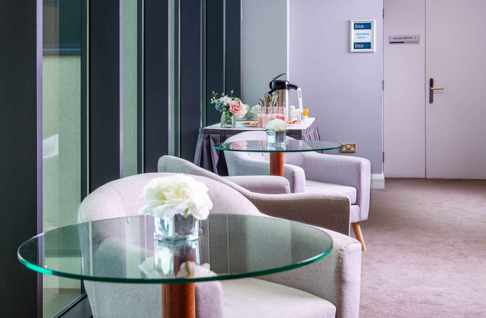 Una habitación de hotel moderna con una mesa de vidrio, dos sillones grises, una mesita de noche con un jarrón de flores y una minibar con aperitivos y bebidas.