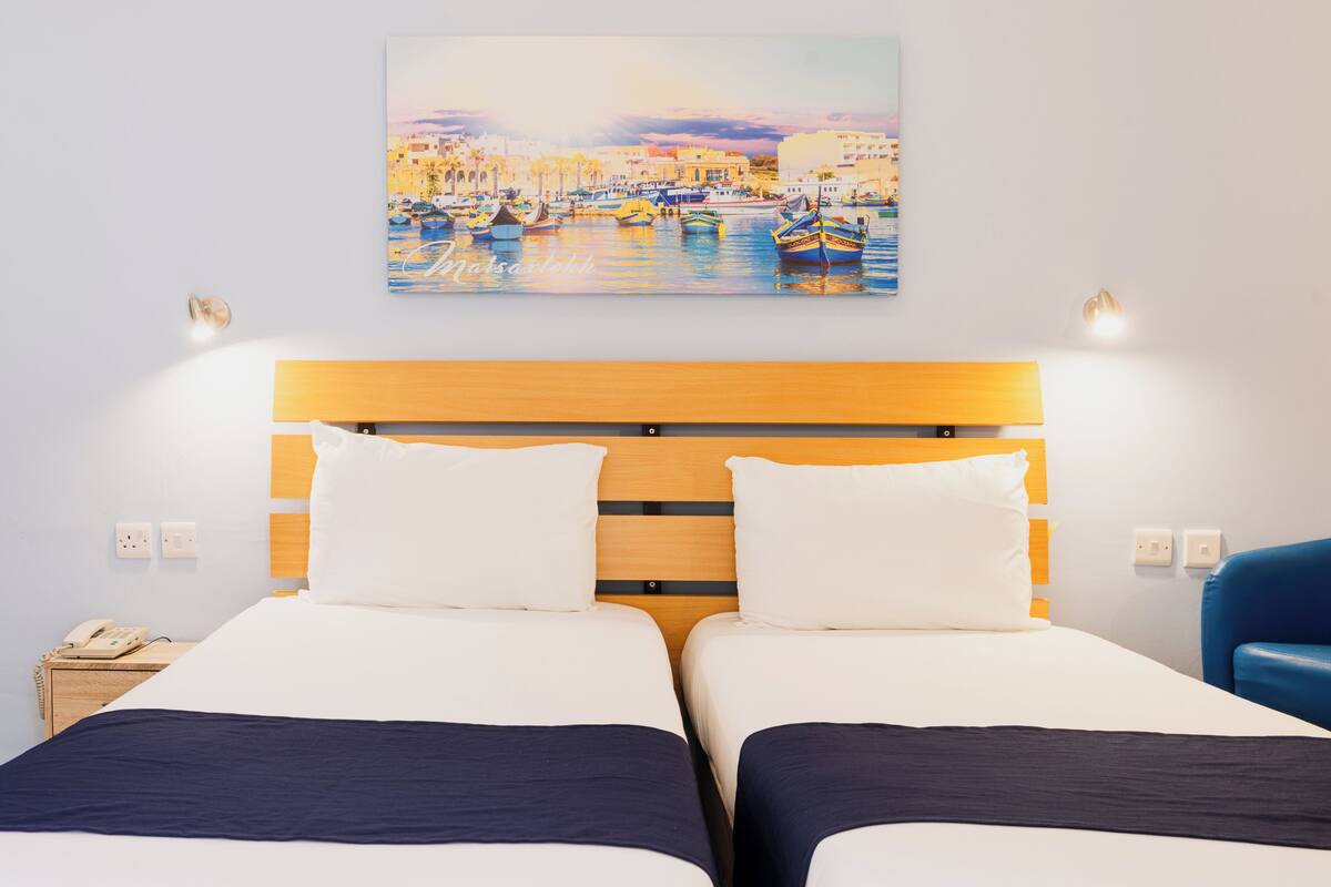 Een hotelkamer met twee bedden, een houten hoofdbord, wit beddengoed, een schilderij van een havenzicht en een blauwe stoel.