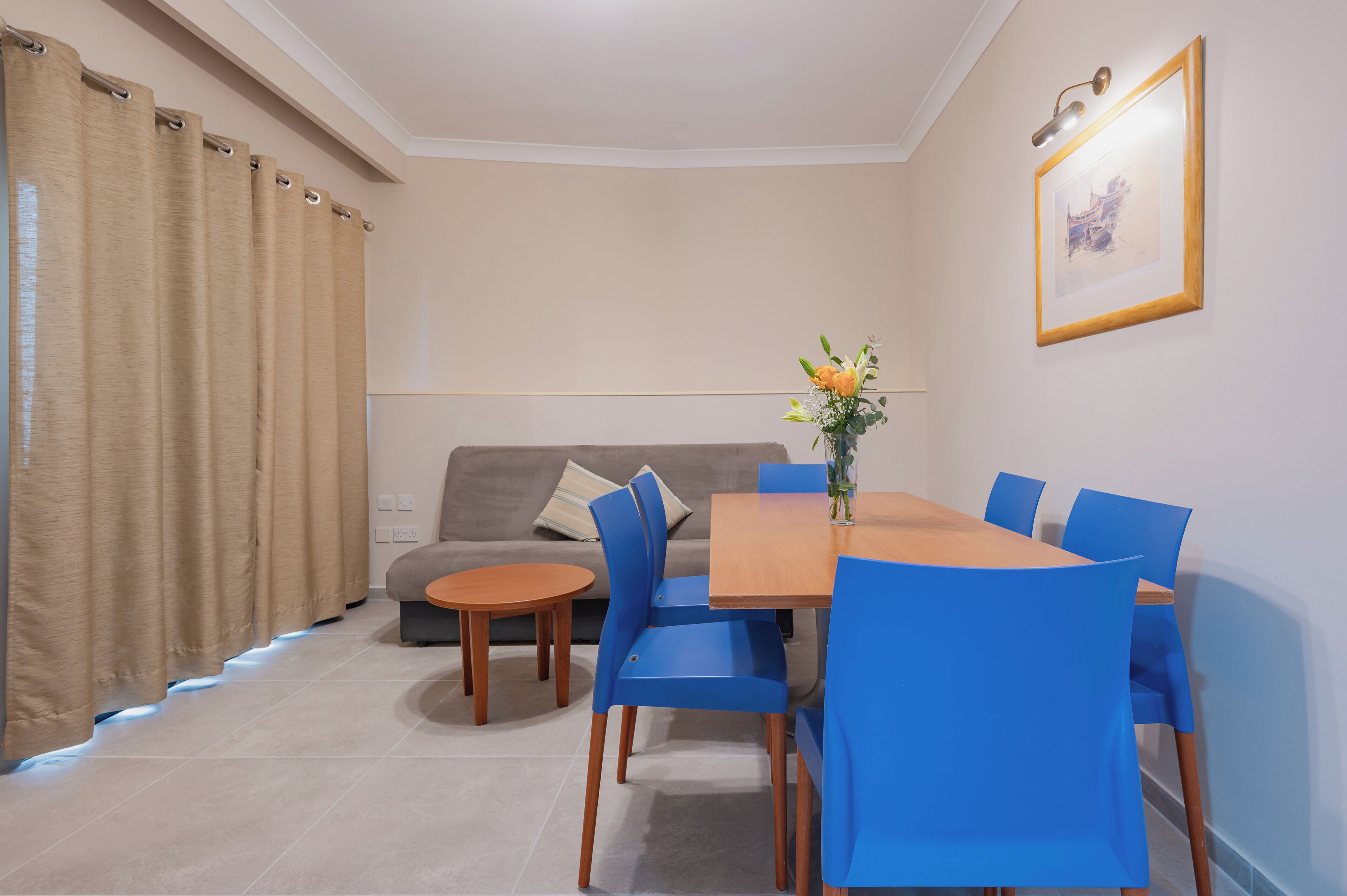 A dining area with a wooden table and blue chairs, a grey sofa with a cushion, and a vase with flowers on the wall.