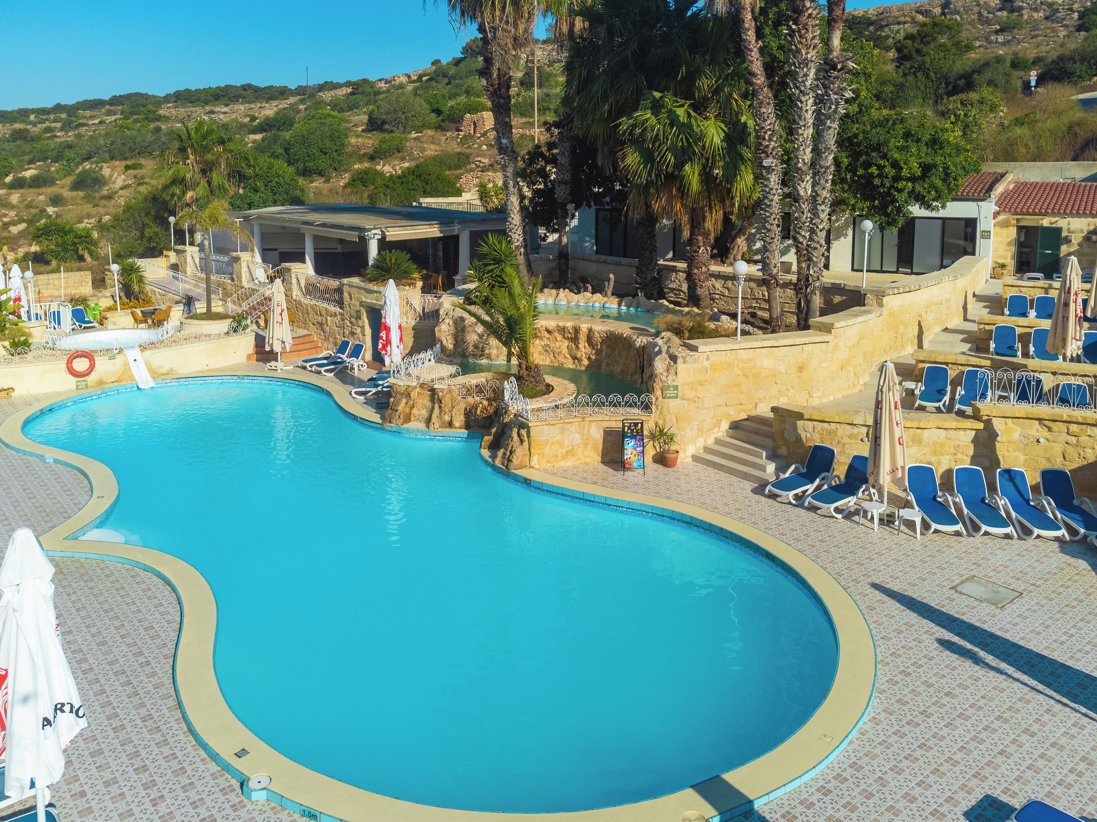 A swimming pool with lounge chairs and umbrellas, surrounded by stone walls and palm trees.