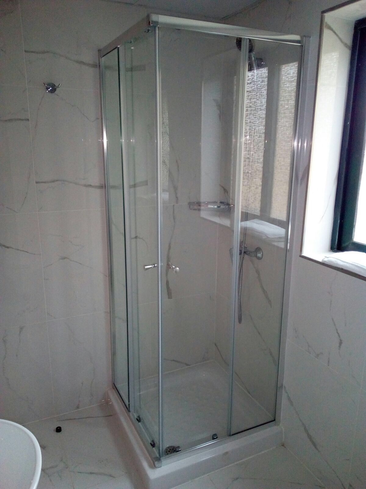 A glass-enclosed shower stall with a white marble floor and walls, a visible showerhead, and a small shelf.