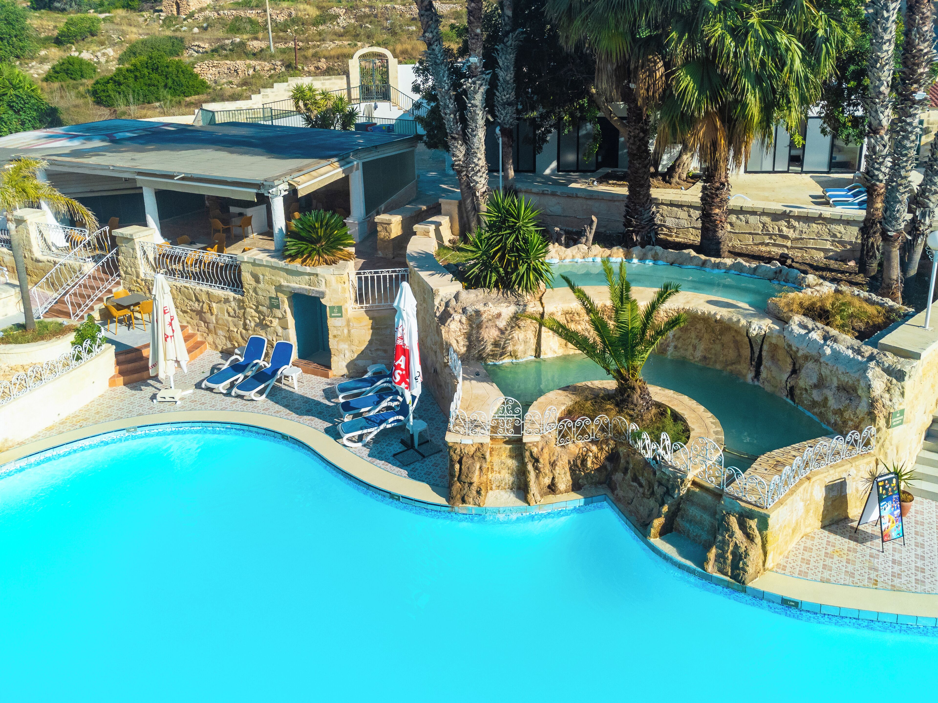 A swimming pool with lounge chairs and umbrellas, surrounded by stone walls and palm trees.