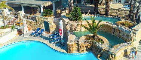 A swimming pool with lounge chairs and umbrellas, surrounded by stone walls and palm trees.