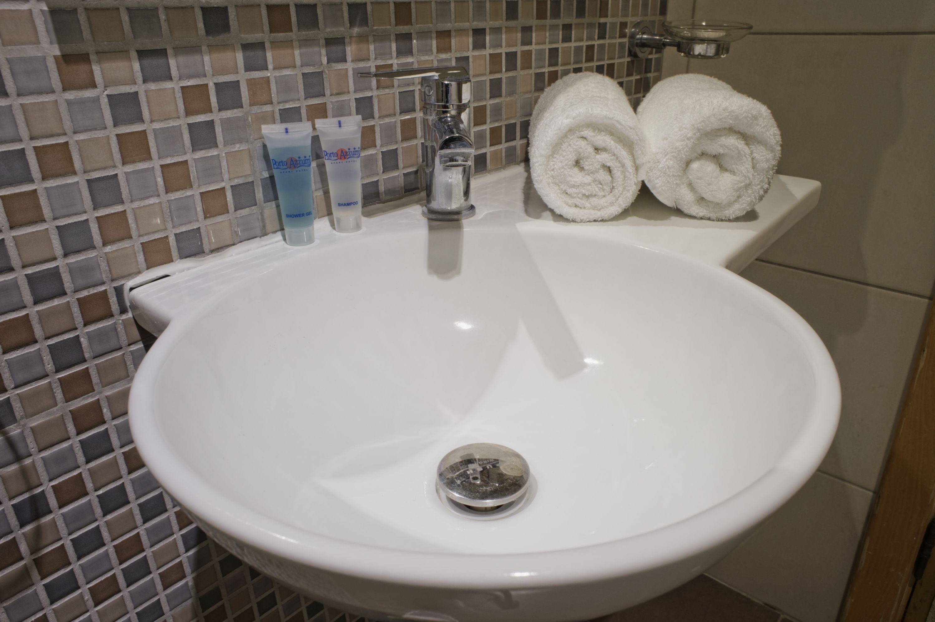 Un lavabo bianco con rubinetteria cromata, due asciugamani bianchi arrotolati e due flaconi di shampoo e balsamo.