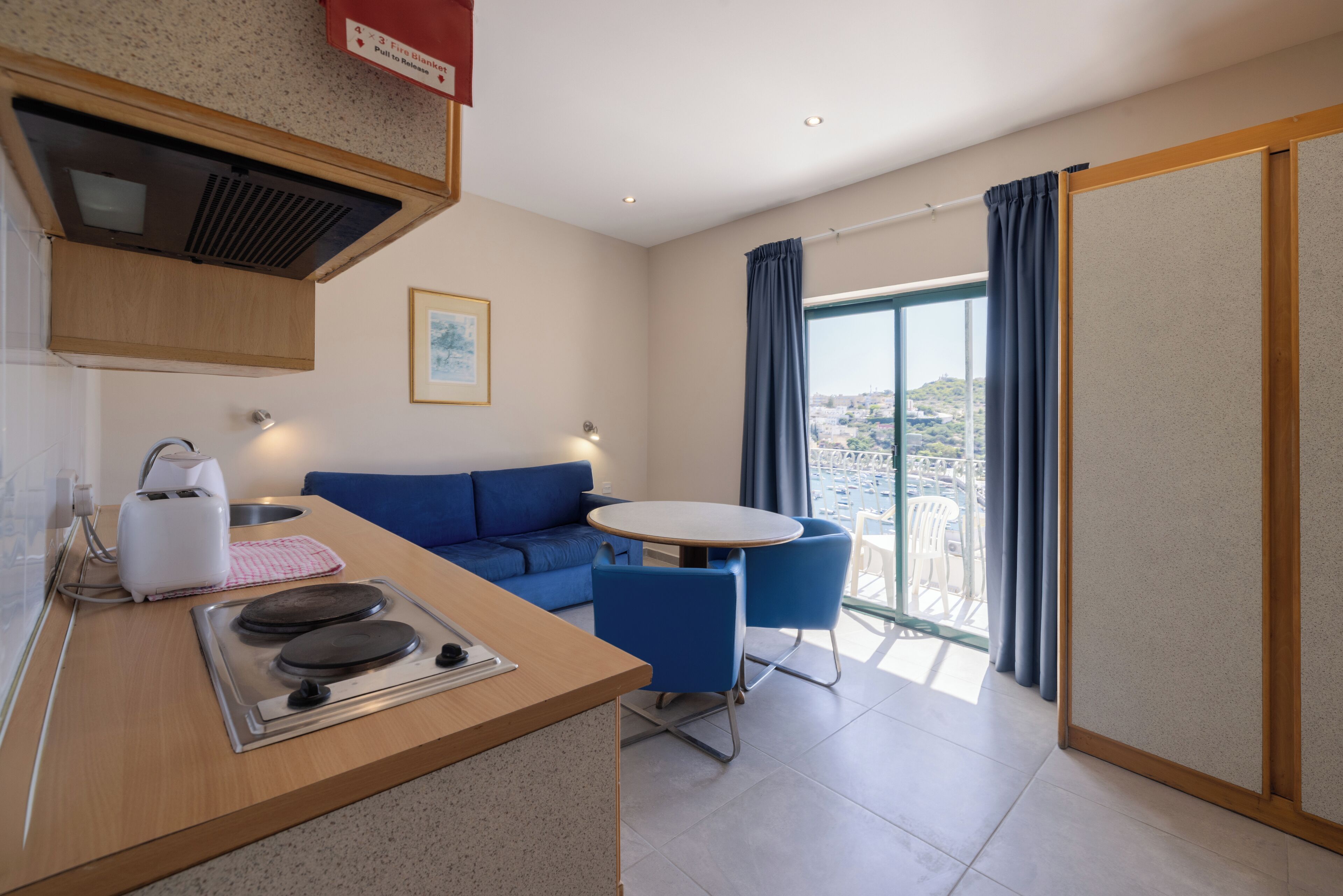 Une cuisine moderne dotée d’une cuisinière intégrée, un coin repas avec une table ronde et des chaises bleues, un canapé et un balcon offrant une vue sur la ville.