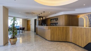 A reception area with a curved wooden counter, a potted plant, and a seating area with tables and chairs. - Xemxija Bay Hotel (St. Paul's Bay)
