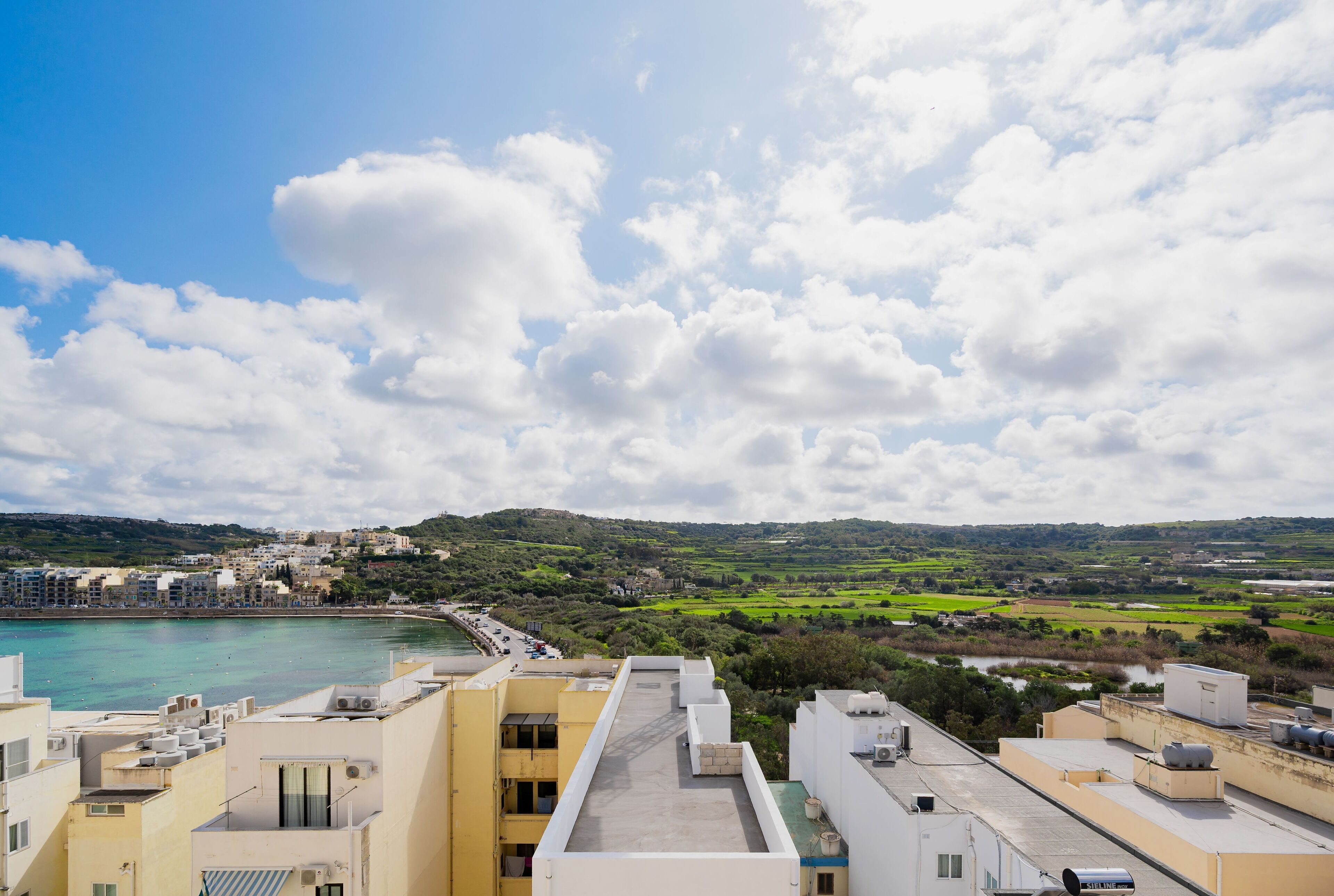 A coastal cityscape with buildings overlooking a bay, featuring a clear sky and distant green hills.