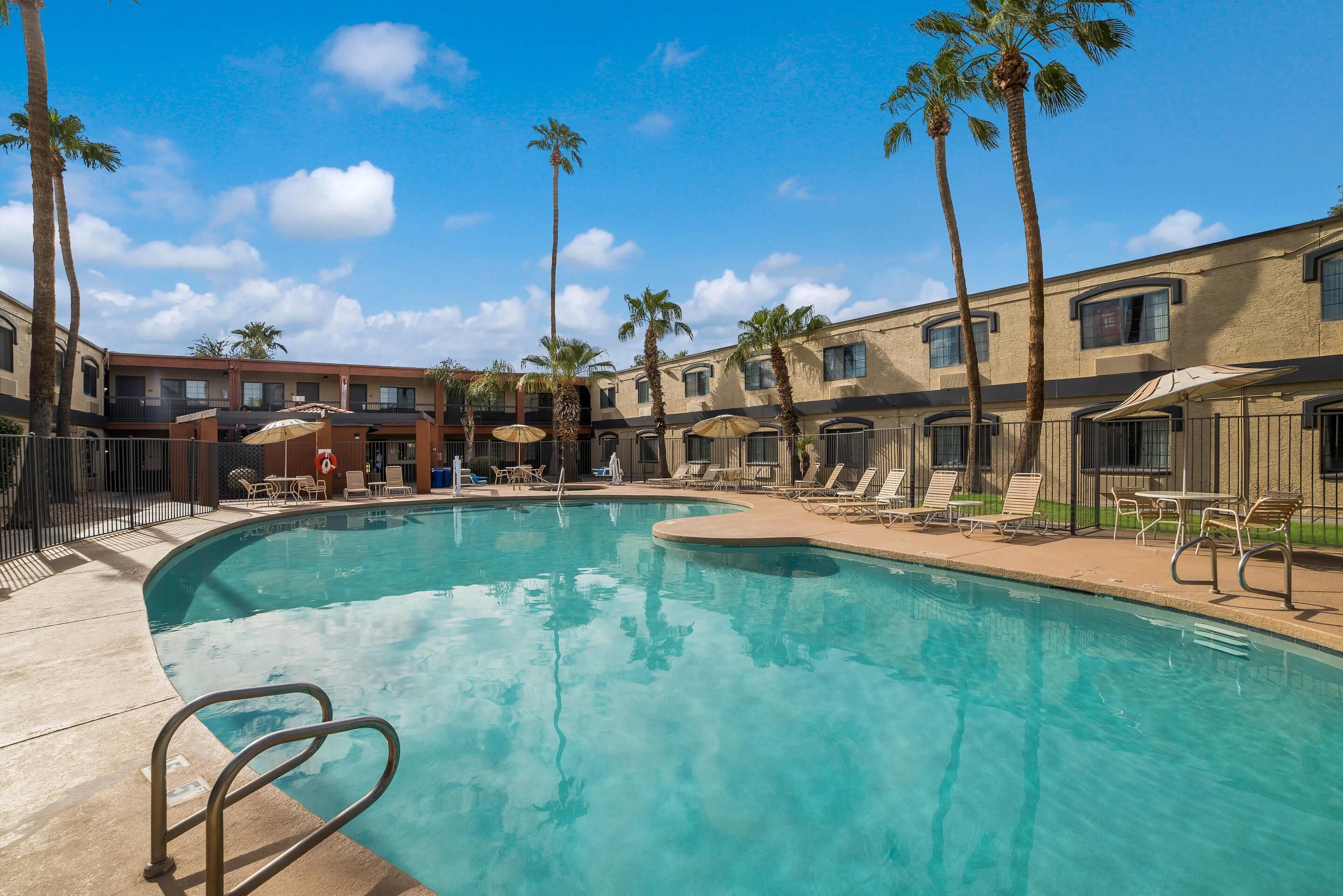 A pool area with lounge chairs and umbrellas, surrounded by palm trees and a building with multiple windows.