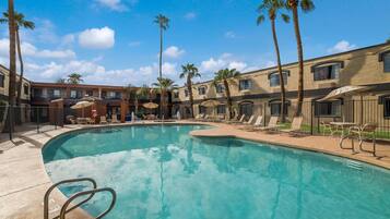 A pool area with lounge chairs and umbrellas, surrounded by palm trees and a building with multiple windows.