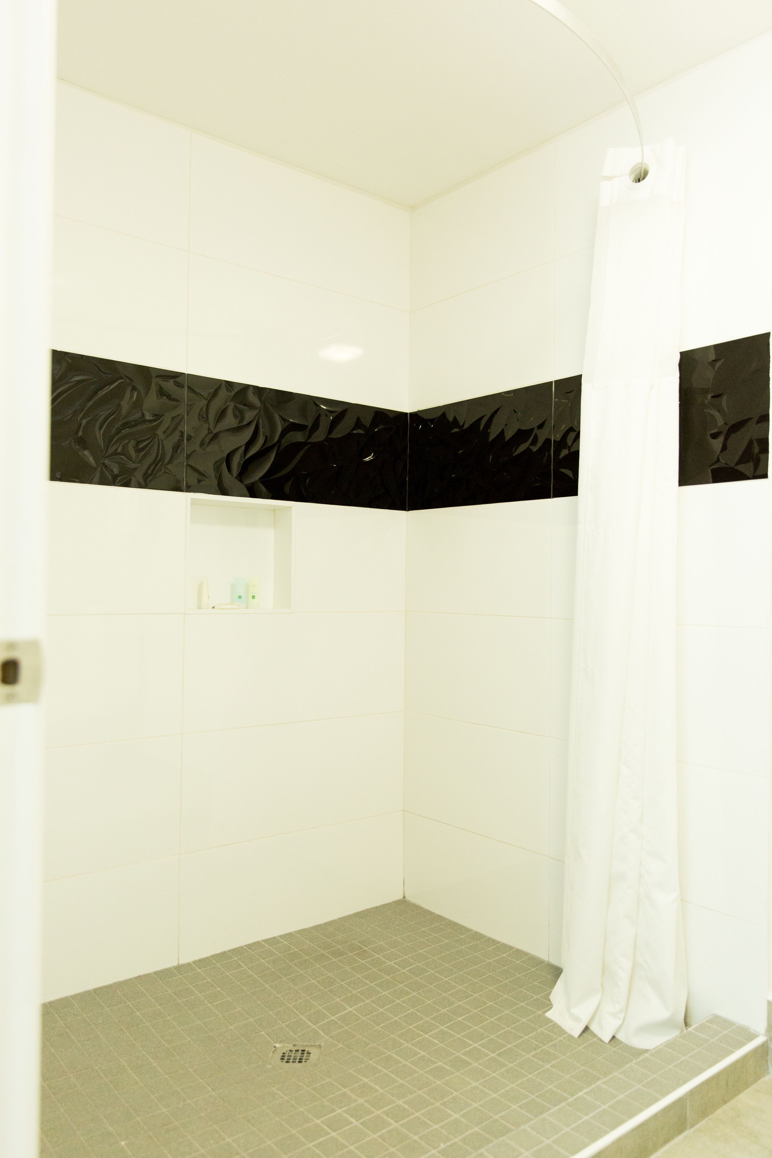 Área de chuveiro com azulejos brancos, uma faixa decorativa preta e um cortina de chuveiro branca.