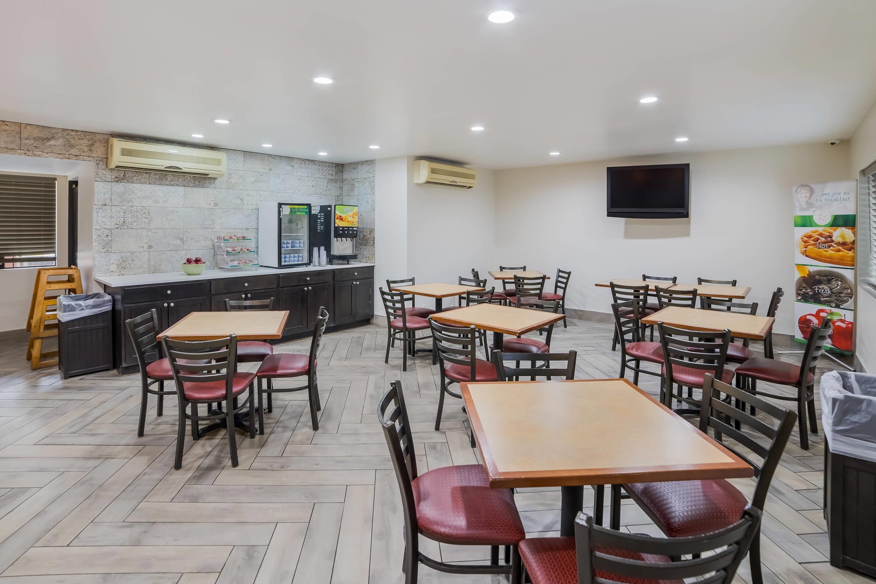 Un restaurante con varias mesas y sillas, una zona de cocina con un refrigerador y una máquina de café, y un televisor de pantalla plana montado en la pared.