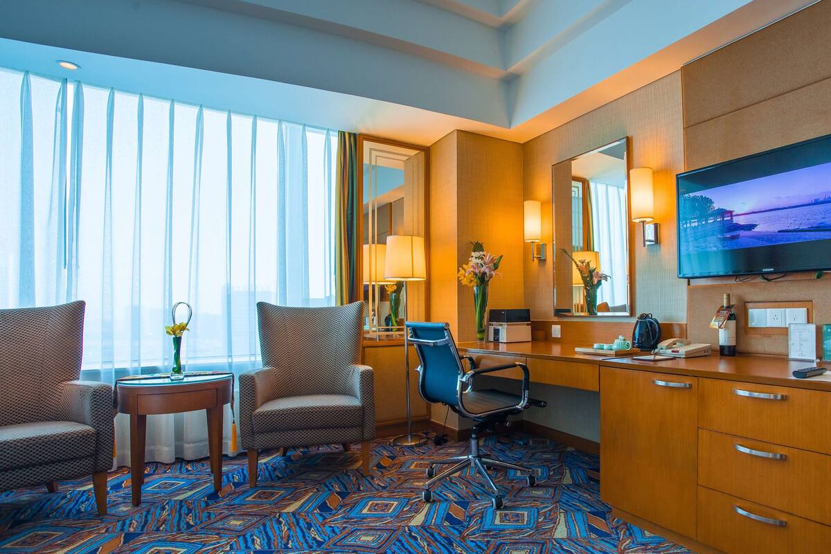 A hotel room with a large window, a desk with a chair, a television, and a minibar.