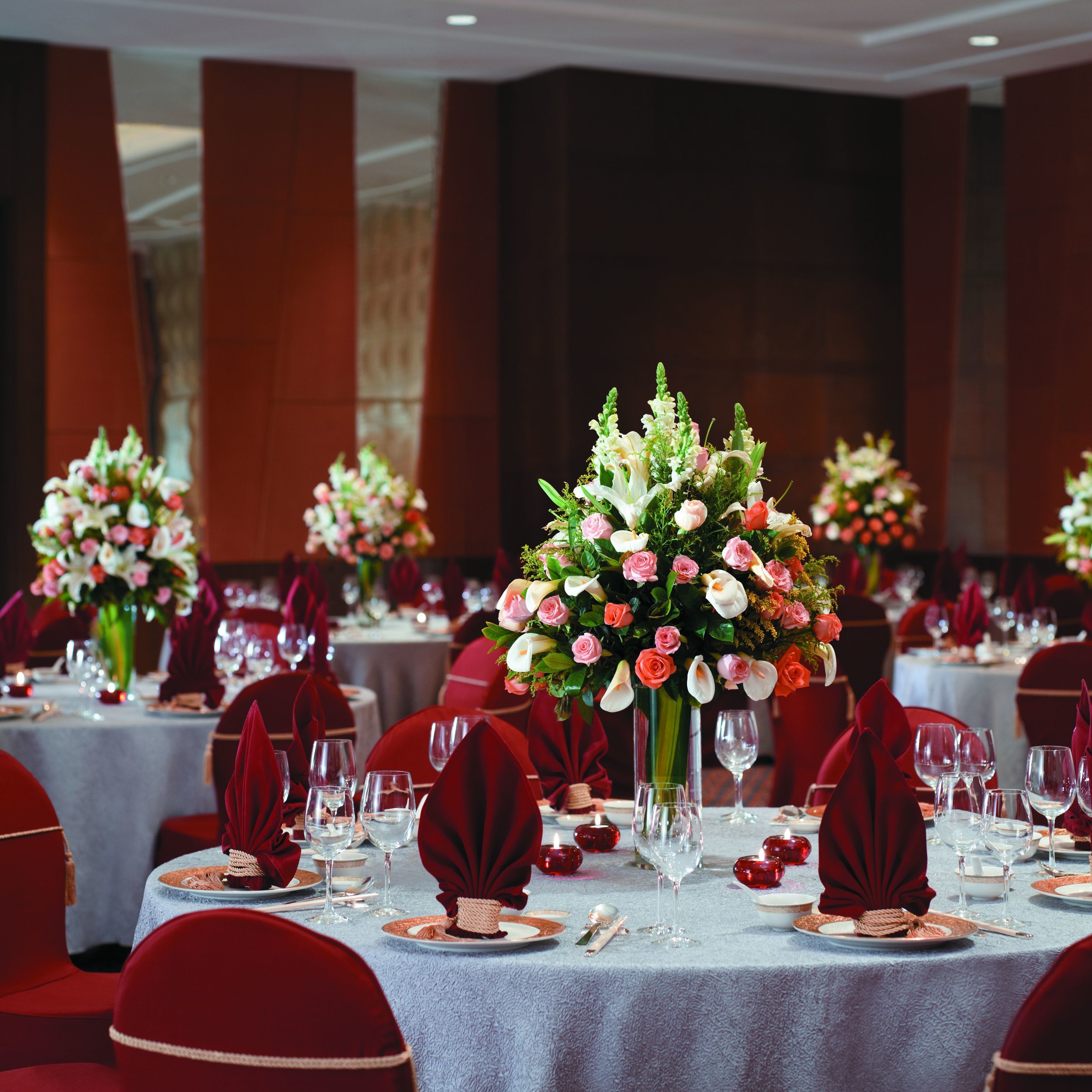 Un salón de banquetes con mesas redondas dispuestas para comer, cada una con un centro de mesa de flores y servilletas rojas.