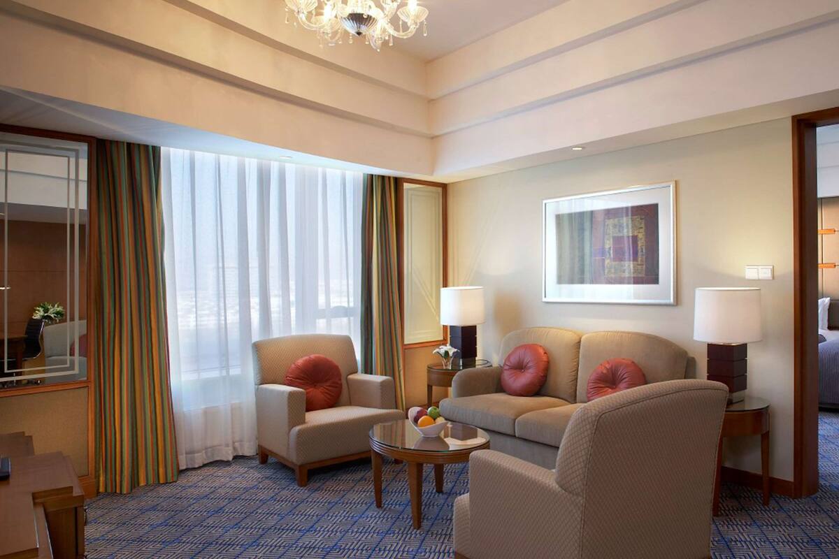 A hotel room with a sofa, armchairs, a small table, a lamp, and a framed picture on the wall.